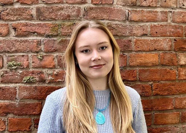 A head and shoulders shot of a young blonde lady stood in front of a brick wall.