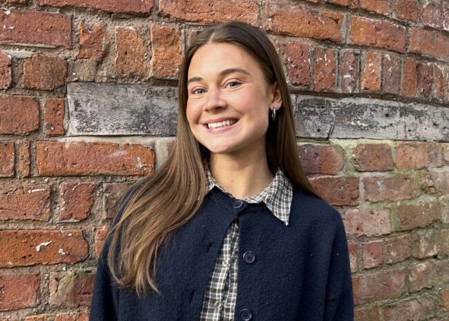 Brunette woman standing against a rustic, red brick wall wearing a navy cardigan.