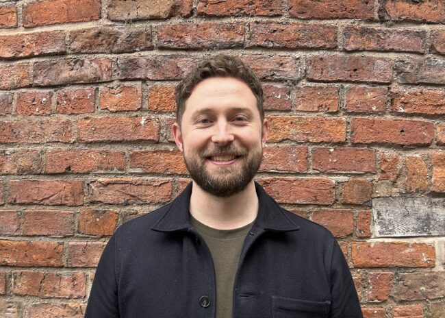 Brunette man with bread, khaki top and navy button jacket standing in front of a brick wall